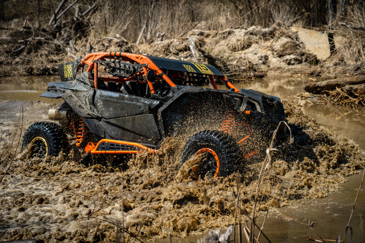 Black and orange powersport vehicle splashes through muddy waters.